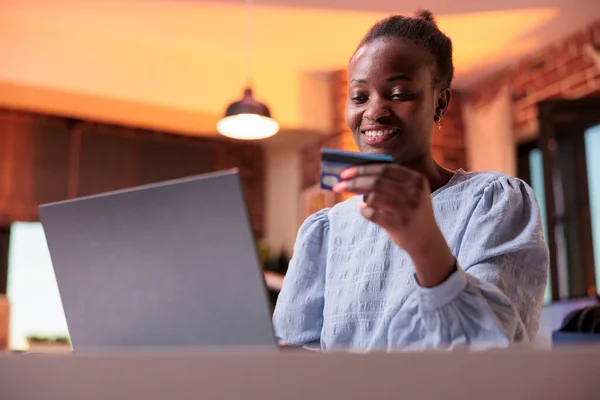 Smiling woman holding debit card and buying products in online store using laptop. Young african american female freelancer searching goods and making purchase in internet shop