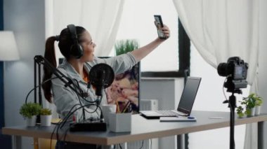 İnternet yıldızı stüdyo masasında oturup selfie çekerken yayın yapıyor. Sosyal medya etkileyicisi modern dokunmatik ekranlı cep telefonuyla fotoğraf çekerken oturma odasında oturuyor.