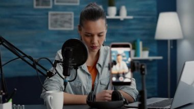 Yaratıcı dijital içerik yaratıcısı akıllı telefonuyla video blogu çekerken stüdyo masasında oturuyor. Mutlu gülümseyen genç sosyal medya etkileyicisi stüdyoda video blogu kaydetmeye hazırlanıyor..