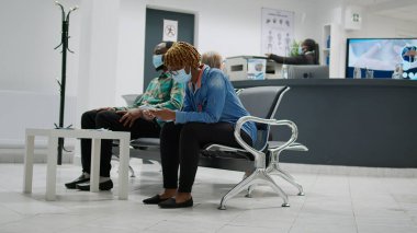 Yüz maskeli çok ırklı bir grup insan bekleme alanında oturuyor, uzman ve önleyici virüs danışmanlarıyla randevuları var. Koronavirüs salgını sırasında hastane resepsiyonu. Üçayak atışı.