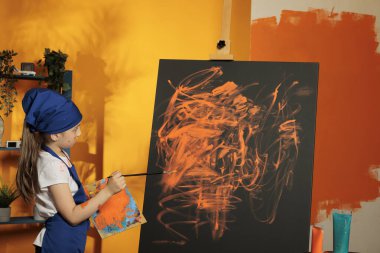 Young kid using orange aquarelle to paint artwork on canvas with easel, doing artistic practice to create masterpiece design with watercolor palette and paintbrush. Painting creation with wet dye.