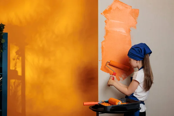 Small kid using orange paint on walls, painting apartment room with roller paintbrush at home. Little girl having fun with housework redecoration and renovation, using brush tools.