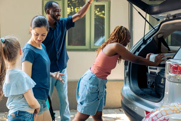 Diverse people loading luggage in trunk of automobile, travelling on summer holiday. Mother with child and diverse friends putting suitcase bags in vehicle, travelling on vacation road trip.