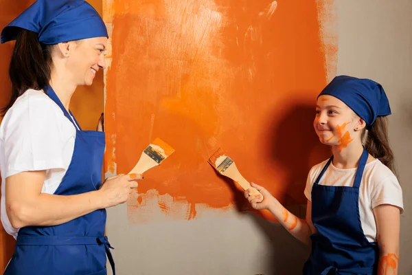 Happy people using brush to paint walls with orange color and renovating tools, having fun with messy redecoration work. Adult with little messy girl decorating apartment interior with paintwork.