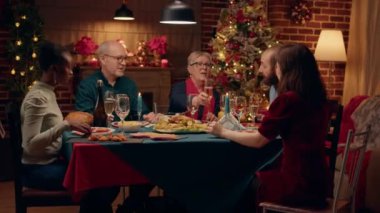 Happy family members toasting champagne while sitting at Christmas dinner table at home. Festive diverse people enjoying reunion while celebrating winter holiday inside Xmas decorated cozy apartment.