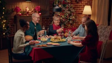 Laughing diverse people gathered around Christmas dinner table clinking wine glasses. Happy smiling family members celebrating traditional winter holiday inside cozy apartment with Xmas setting.