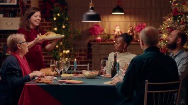 Beautiful wife bringing main course to Christmas dinner table at home. Joyful multiethnic family members celebrating winter holiday inside cozy apartment with traditional Xmas decorations. Tripod shot
