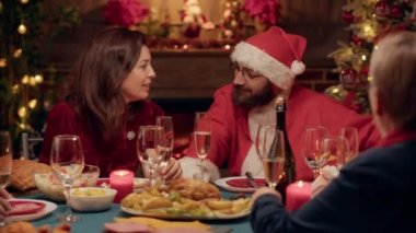 Festive married couple discussing at Christmas dinner while enjoying time with loved ones. Young adult person disguised as Santa Claus celebrating winter holiday with family members at home.