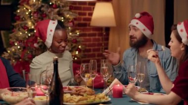 Festive husband talking with african american woman while clinking champagne glasses at Christmas dinner. Happy diverse people wearing traditional hats celebrating winter holiday.