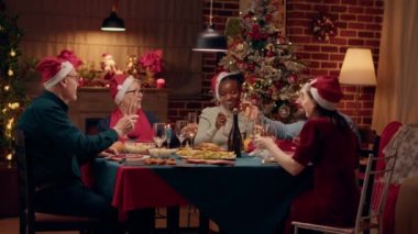 Cheerful festive people singing traditional songs while enjoying Christmas dinner together at home. Happy family members drinking champagne while celebrating winter holiday. Tripod shot