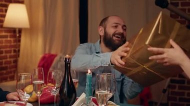 Multiethnic people gathered in living room to celebrate winter holiday while giving each other gifts. Festive family members sitting at Christmas dinner table while exchanging presents at home.