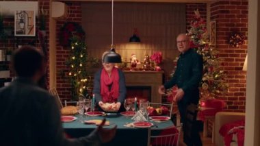 Festive senior couple welcoming joyful diverse people at Christmas family dinner table in living room. Cheerful people gathering to celebrate winter traditional holiday inside Xmas decorated apartment