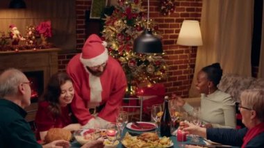 Funny husband wearing Santa Claus costume offering gifts to family while celebrating winter holiday at home. Wholesome man disguised as Santa Claus giving festive family presents at Christmas dinner.