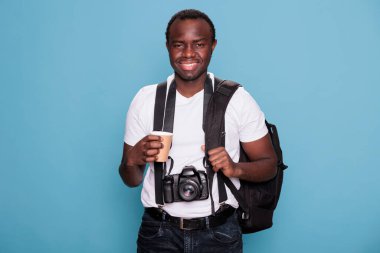 Tourist with professional camera and travel backpack ready for holiday journey on citybreak. African american photographer with DSLR photo device and travel roadtrip bag standing on blue background.
