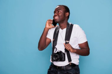 Happy professional photographer having DSLR camera talking on phone while standing and smiling on blue background. Joyful photography enthusiast talking on mobile phone while having photo device.