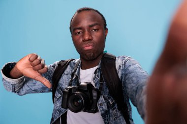 African american photography enthusiast with DSLR device showing thumbs down hand gesture at camera while standing on blue background. Photographer with photo device giving disapprove finger symbol.