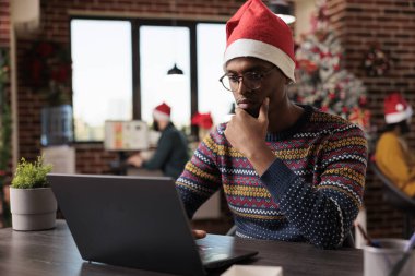 African american man working on laptop in office, using computer in festive workplace decorated with christmas tree and holiday ornaments. Male worker at company job during winter season.