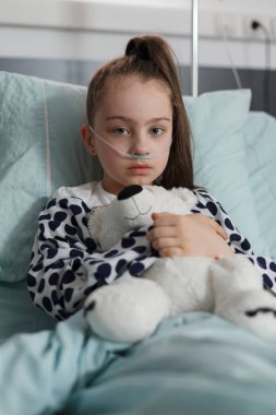 Sick kid wearing oxygen tube while sitting in patient bed having plush bear toy. Under treatment ill little girl resting in pediatric healthcare facility while holding teddybear.