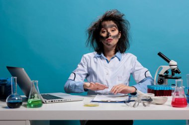 Mad silly looking scientist sitting at desk in laboratory with messy hair and dirty face after failed chemical experiment. Maniac goofy chemist with exploded look and smokey face looking at camera.