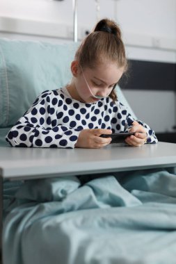 Hospitalized ill kid playing games on modern mobile phone while resting inside hospital pediatrics ward. Sick girl under treatment resting on patient bed while watching cartoons on smartphone.