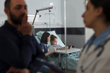 Hospitalized ill little kid watching cartoons on mobile phone while resting in hospital pediatric ward. Sick girl playing games on smartphone with mother while father talking with expert pediatrist.