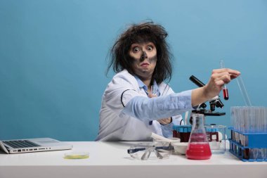 Dumb looking crazy chemist with wacky hairstyle and dirty face sitting at desk in laboratory handling glass test tubes. Mad goofy scientist holding glass beaker while looking silly at camera.