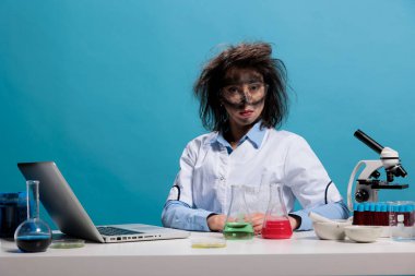 Foolish lab worker with wacky hair and dirty face sitting at desk on blue background while looking at camera. Silly looking crazy chemist with messy hairstyle after failed experiment.