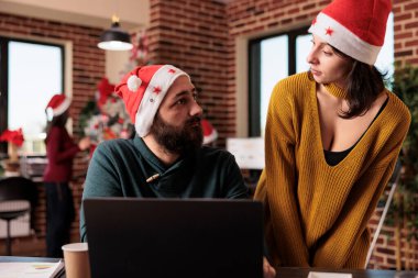 Festive coworkers doing teamwork in startup office with christmas tree and lights, using laptop to work on business. Man and woman with santa hat at job with holiday decorations and ornaments.