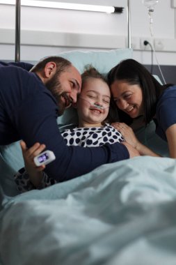 Loving parents hugging sick daughter resting in healthcare clinic pediatrics ward. Happy ill little girl hugged by caring mother and father while sitting with her on patient bed.