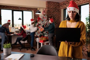 Woman in santa hat using laptop in festive office, working on business in workplace with christmas tree and ornaments. Colleagues decorating job space with seasonal xmas lights.