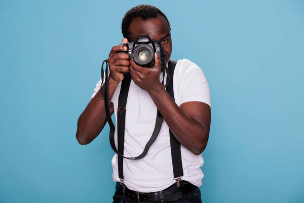 African american photography enthusiast taking photo while standing on blue background. Confident professional photographer having DSLR camera taking picture. Studio shot.