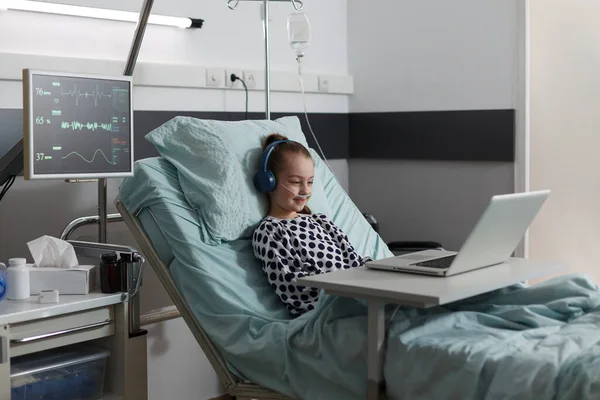 Happy sick little girl under treatment sitting on patient bed with laptop while watching cartoons inside hospital pediatrics ward room. Ill kid having oxygen tube enjoying funny videos on computer.