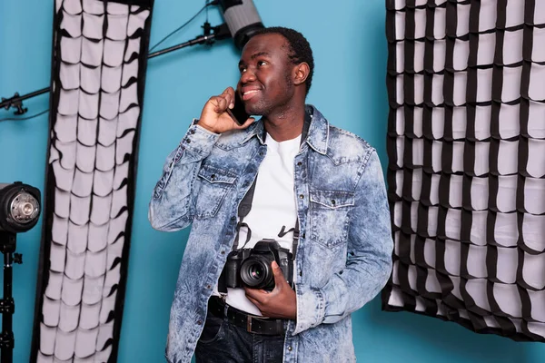 Professional photographer talking on smartphone while standing in studio having softboxes and production equipment. Creative video producer on blue background with spotlight while having a phone call.