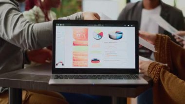 Multiethnic group of coworkers analyzing business charts and infographics data on laptop software. People doing teamwork collaboration to create paper reviews and professional report. Close up.