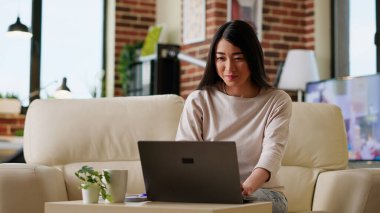 Smiling heartily woman at home working remotely on laptop while sitting on couchin living room. Joyful young adult freelancer doing remote work on modern portable computer while at home.