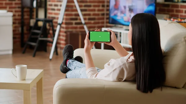 Relaxed person with modern touchscreen mobile phone having chroma key isolated display. Woman sitting on sofa while holding smartphone with green screen mockup template background. Tripod shot