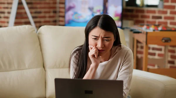 Worried asian person looking confused at computer screen while doing remote work from home. Stressed woman working remotely on laptop while trying to understand job assignment