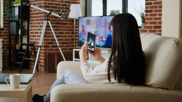Childish young adult person enjoying mobile gaming while sitting on sofa inside apartment. Playful asian woman playing space shooter game on smartphone device while working remotely from home.