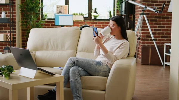 Joyful young adult person texting with friend while sitting on sofa inside apartment. Smiling heartily woman watching funny content on smartphone while drinking coffee inside living room on couch.