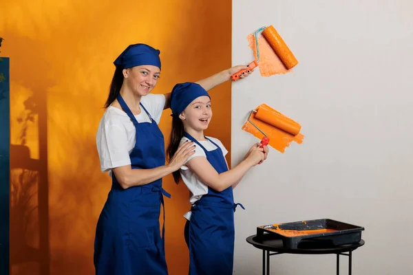 Portrait of family using orange paint to change walls color in apartment room, painting with roller brush to renovate house. Woman with small kid doing diy home redecoration together.