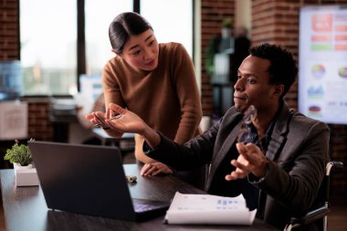 African american wheelchair user working with asian woman on business project in disability friendly office. Paralyzed man with chronic impairment doing teamwork with colleague.
