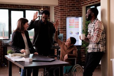 Female manager in wheelchair and group of people laughing on job break, having fun in disability friendly office. Woman with chronic health condition chatting with cheerful colleagues.