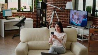 Attractive woman chatting on smartphone while sitting on sofa at home and smiling heartily at camera. Happy young adult person sending messages on modern mobile phone while working remotely.