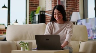 Smiling heartily woman at home working remotely on laptop while sitting on couchin living room. Joyful young adult freelancer doing remote work on modern portable computer while at home.