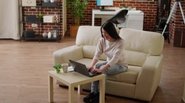 Çekici Asyalı serbest yazar modern taşınabilir bilgisayarda uzaktan kumandayla çalışıyor. Odaklanmış, yürekten gülümseyen bir yetişkin evdeki koltukta oturmuş dizüstü bilgisayarla çalışıyor..