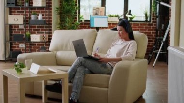 Focused working woman wearing wireless headphones taking notes while doing remote work at home sitting on sofa. Serious looking student attending online class on modern laptop.