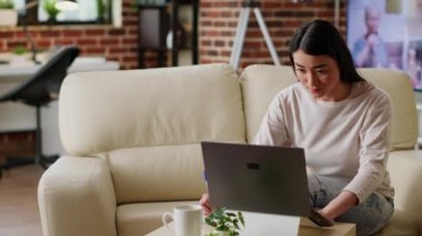 Focused student taking notes while attending online class on laptop at home. Young adult woman writing on notebook while being attentive to modern portable computer in living room.