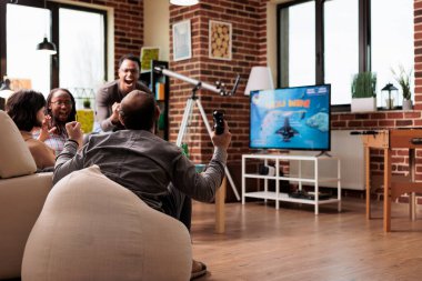 Multiethnic group of friends celebrating console videogame space simulator victory at home. Diverse people sitting on sofa in living room while enjoying games on electronic entertainment device.
