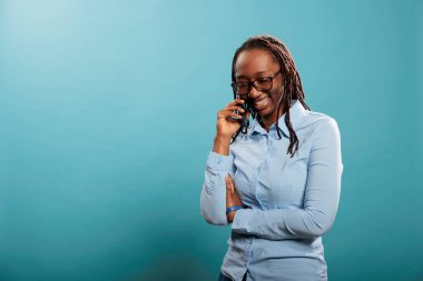 Happy joyful young woman smiling heartily while having a phone call conversation on blue background. Cheerful adult person talking with friend on touchscreen cellphone device.