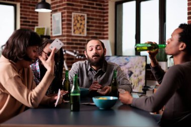 Playful multiethnic friends at home playing card games together while having snacks and beverages. Diverse group of people sitting at table in living room while relaxing with fun leisure activity.